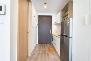 A narrow hallway with a wooden floor leading to a dark door. A modern kitchen with a stainless steel refrigerator and white cabinets is visible to the right.
