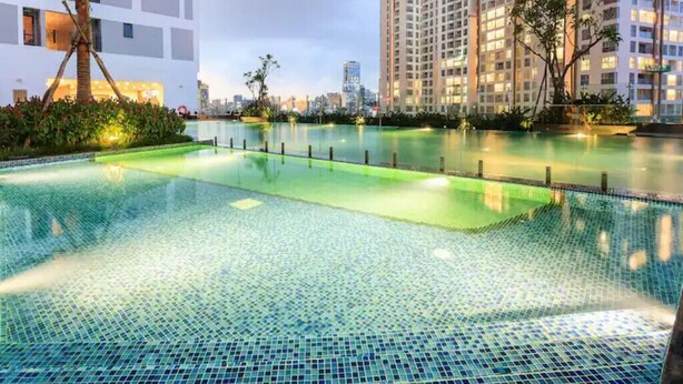A swimming pool with a tiled floor, surrounded by greenery and modern buildings. - Cozy 2 Bedroom Apartment, Free Pool Near Bui Vien (Thành phố Hồ Chí Minh)