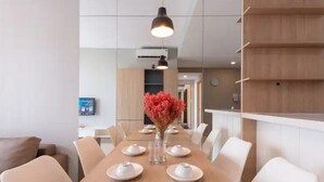 A dining area with a long wooden table set for four, white chairs, a vase with red flowers, and pendant lighting. - Cozy 2 Bedroom Apartment, Free Pool Near Bui Vien (Thành phố Hồ Chí Minh)