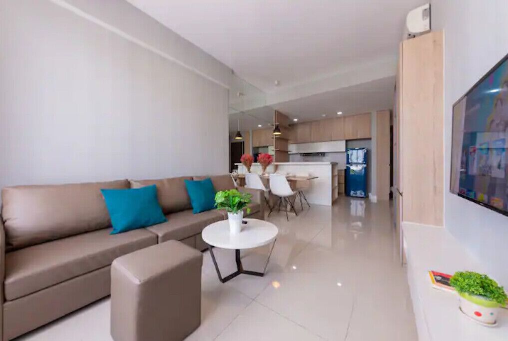 A modern living room with a sofa, a coffee table, and a TV. There is a kitchen area in the background with cabinets and a refrigerator.