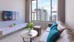 A modern living room with a large window, a flat-screen TV mounted on the wall, a white round coffee table, a brown leather sofa with teal cushions, and a small potted plant. - Cozy 2 Bedroom Apartment, Free Pool Near Bui Vien (Thành phố Hồ Chí Minh)