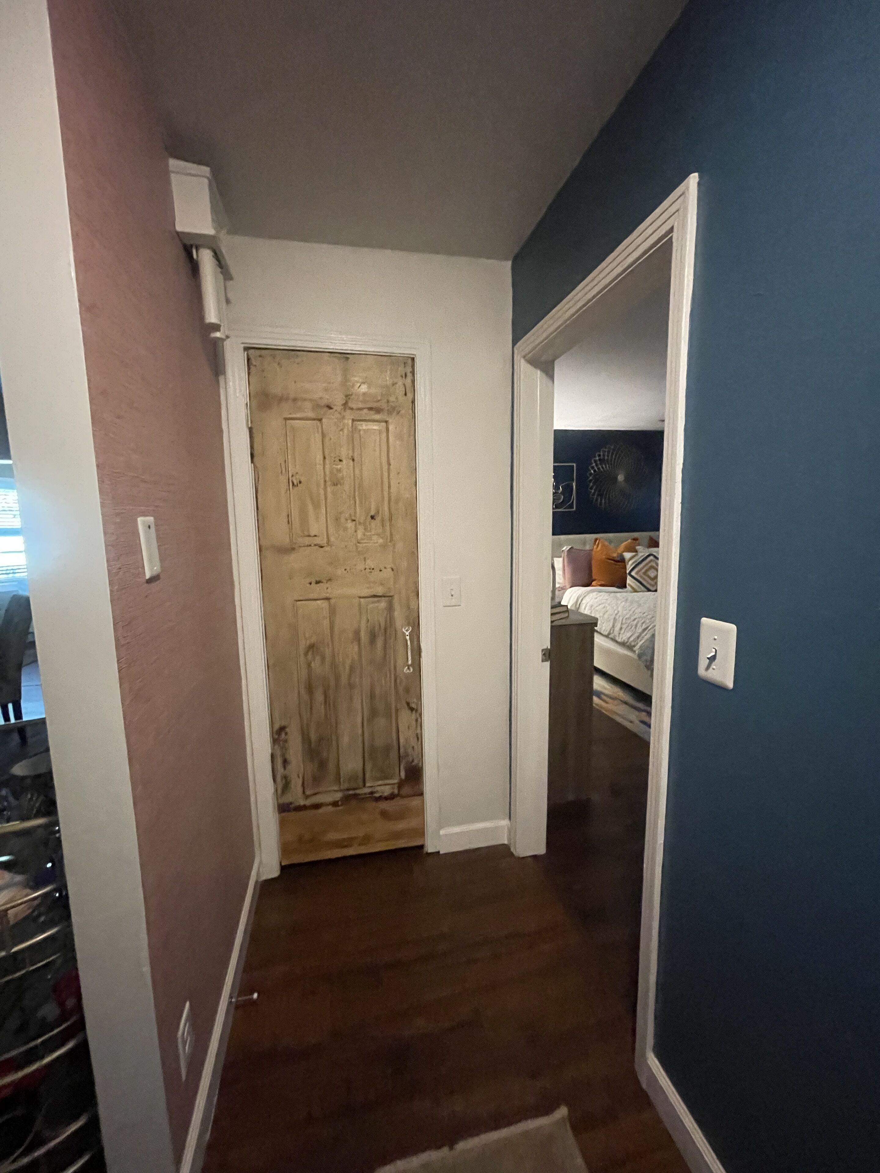 Un pasillo con una puerta de madera, una pared azul y una cama visible en la habitación contigua.