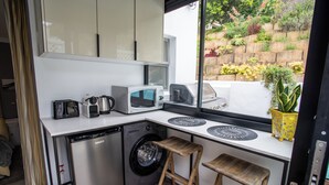 A compact kitchen with a washing machine, a microwave, a toaster, and a coffee maker. There are two wooden stools and a potted plant on the counter. - Camps Bay Echo Garden Apartment (Cape Town)