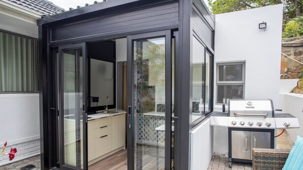 A modern outdoor kitchen with a sliding glass door, a barbecue grill, and a wicker sofa. - Camps Bay Echo Garden Apartment (Cape Town)