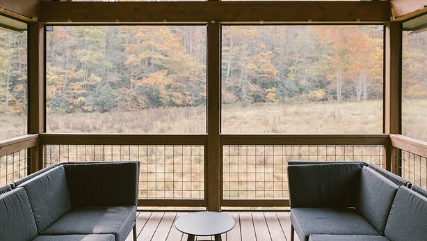 A covered outdoor seating area with two sofas and a small table, large windows offering a view of a forested landscape. - Scandi Cabin on 105 acres: Hot Tub/Sauna/Views/EV, (Lansing)