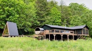 A cabin with a wooden deck and a large cylindrical structure, surrounded by lush greenery. - Scandi Cabin on 105 acres: Hot Tub/Sauna/Views/EV, (Lansing)