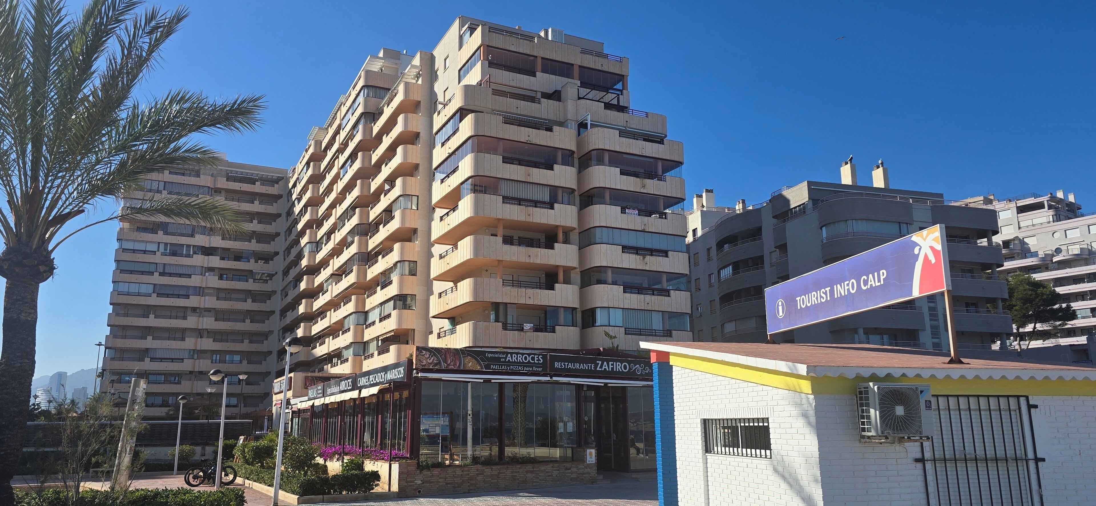 Un edificio residenziale a più piani con balconi, un ristorante chiamato 'ZAFIRO' al piano terra e un cartello con scritto 'TOURIST INFO CALP'.