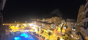 Vista noturna de uma cidade costeira com edifícios iluminados, piscina e palmeiras.