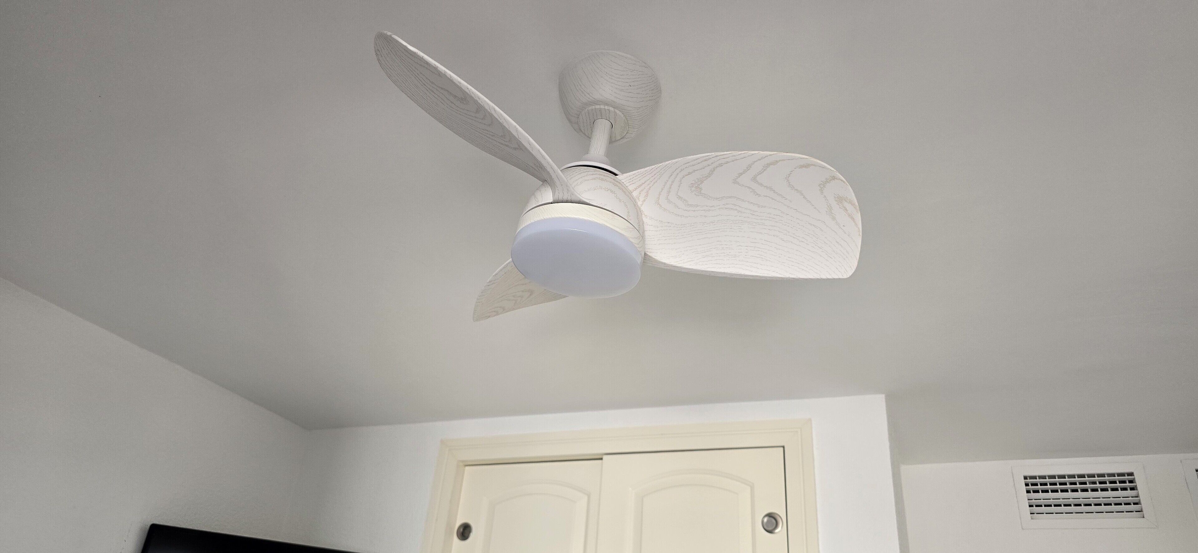 Un ventilateur de plafond avec un luminaire, dans une pièce aux murs blancs et dotée d’une porte.