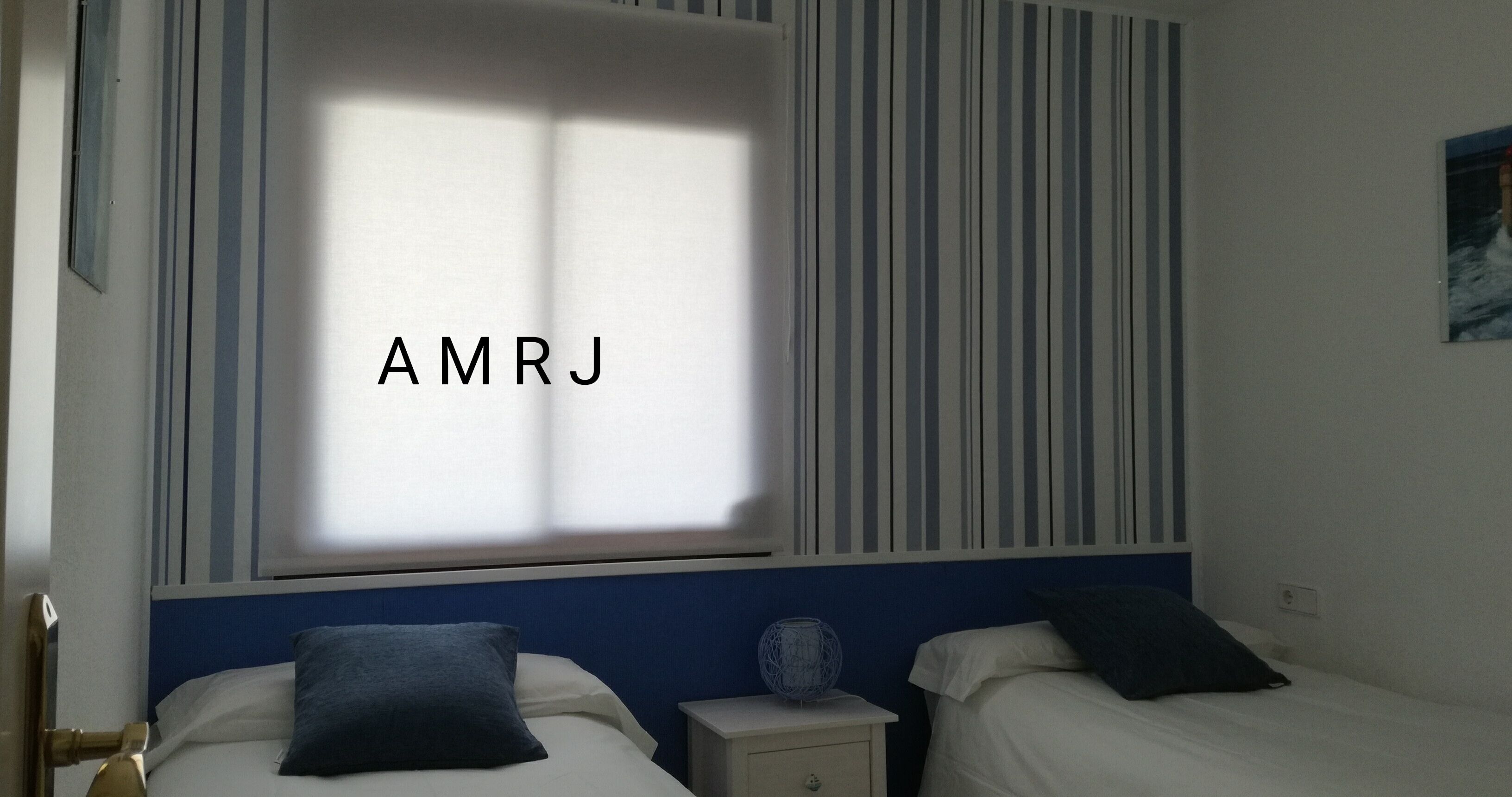 A hotel room with two beds, a window with striped blinds, and the initials "AMRJ" on the window.