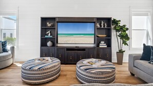 Un salon doté d’un grand téléviseur à écran plat, de deux poufs à motifs, d’un canapé gris et d’une plante en pot.