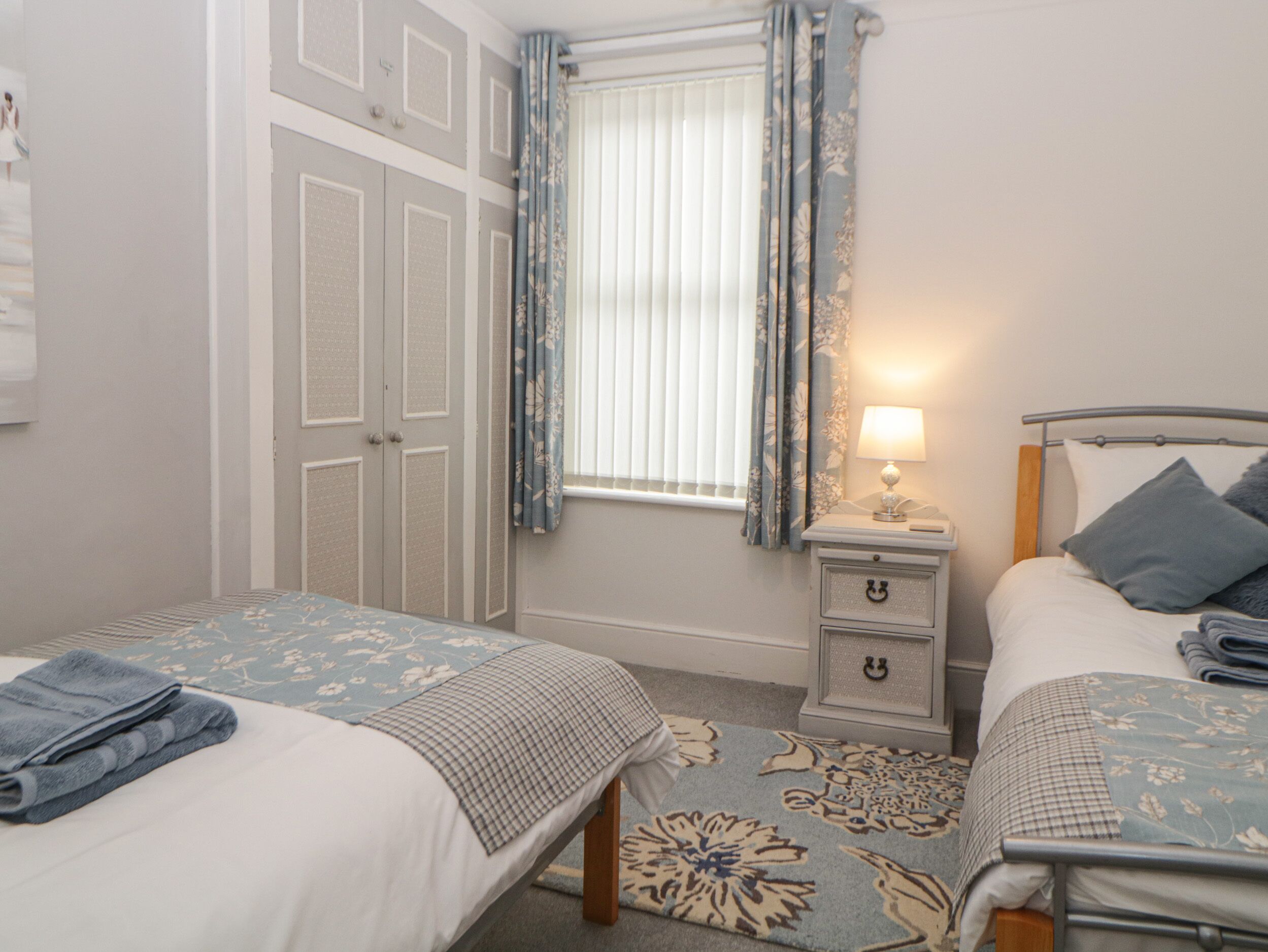A bedroom with two beds, a nightstand, a window with curtains, and a floral carpet.