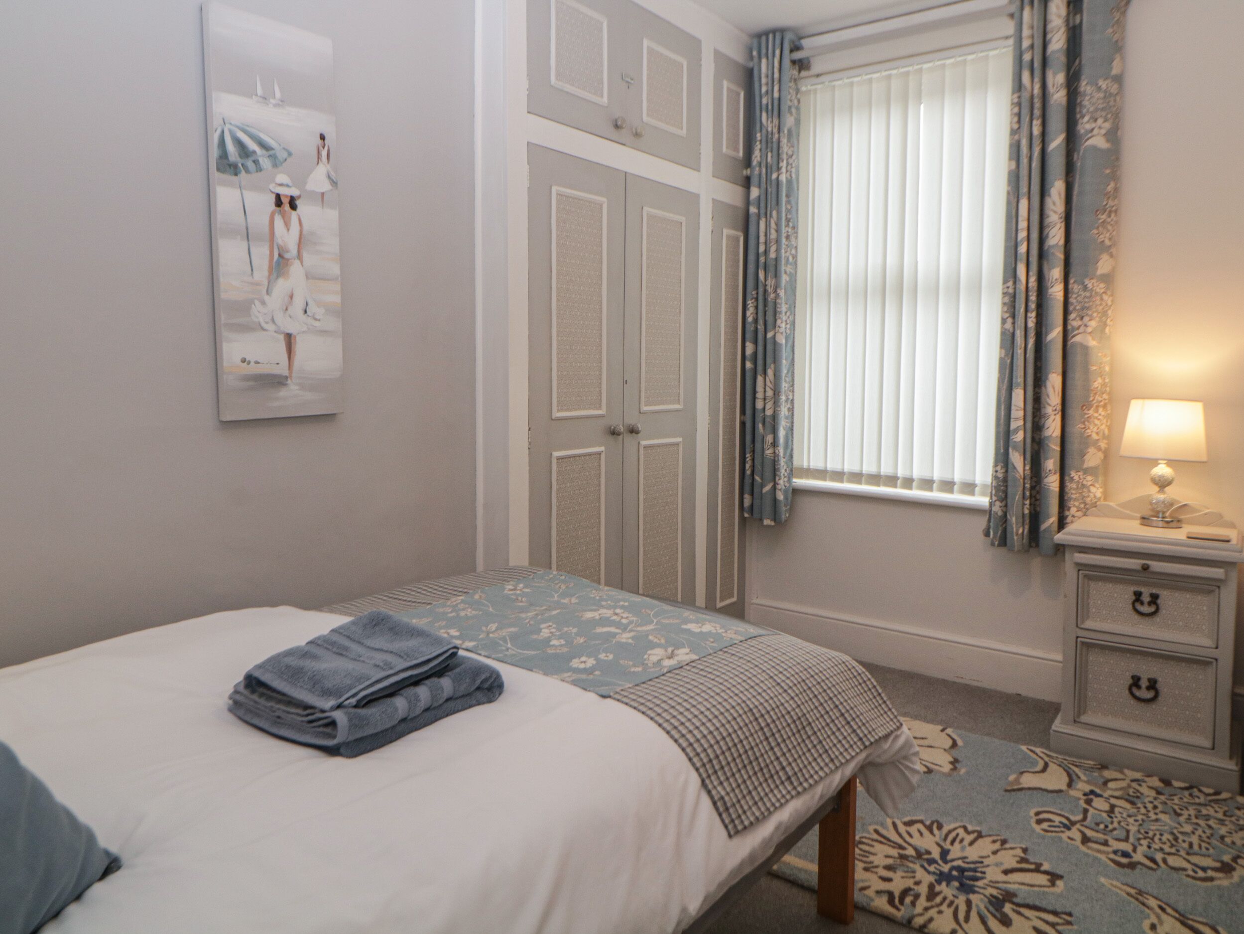 Un dormitorio con cama, una mesita de noche con lámpara, una ventana con cortinas y un cuadro en la pared.
