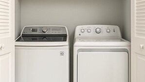 Une buanderie équipée d’un lave-linge et d’un sèche-linge, avec une étagère en bois au-dessus et un mur de couleur gris clair.