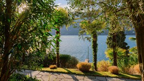 Um jardim arborizado com um caminho de pedra que leva a um corpo d'água, cercado por árvores e arbustos diversos.