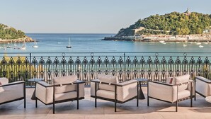 A terrace with outdoor seating overlooking a coastal view with boats and a hill with a statue. - Nobu Hotel San Sebastián (San Sebastián)