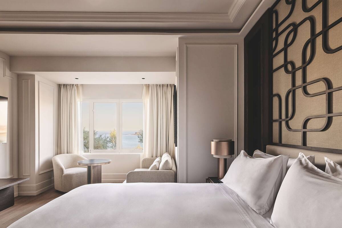 A hotel room with a large bed, a sitting area with a chair and a small table, a view of the outdoors through a window, and a decorative wall panel.
