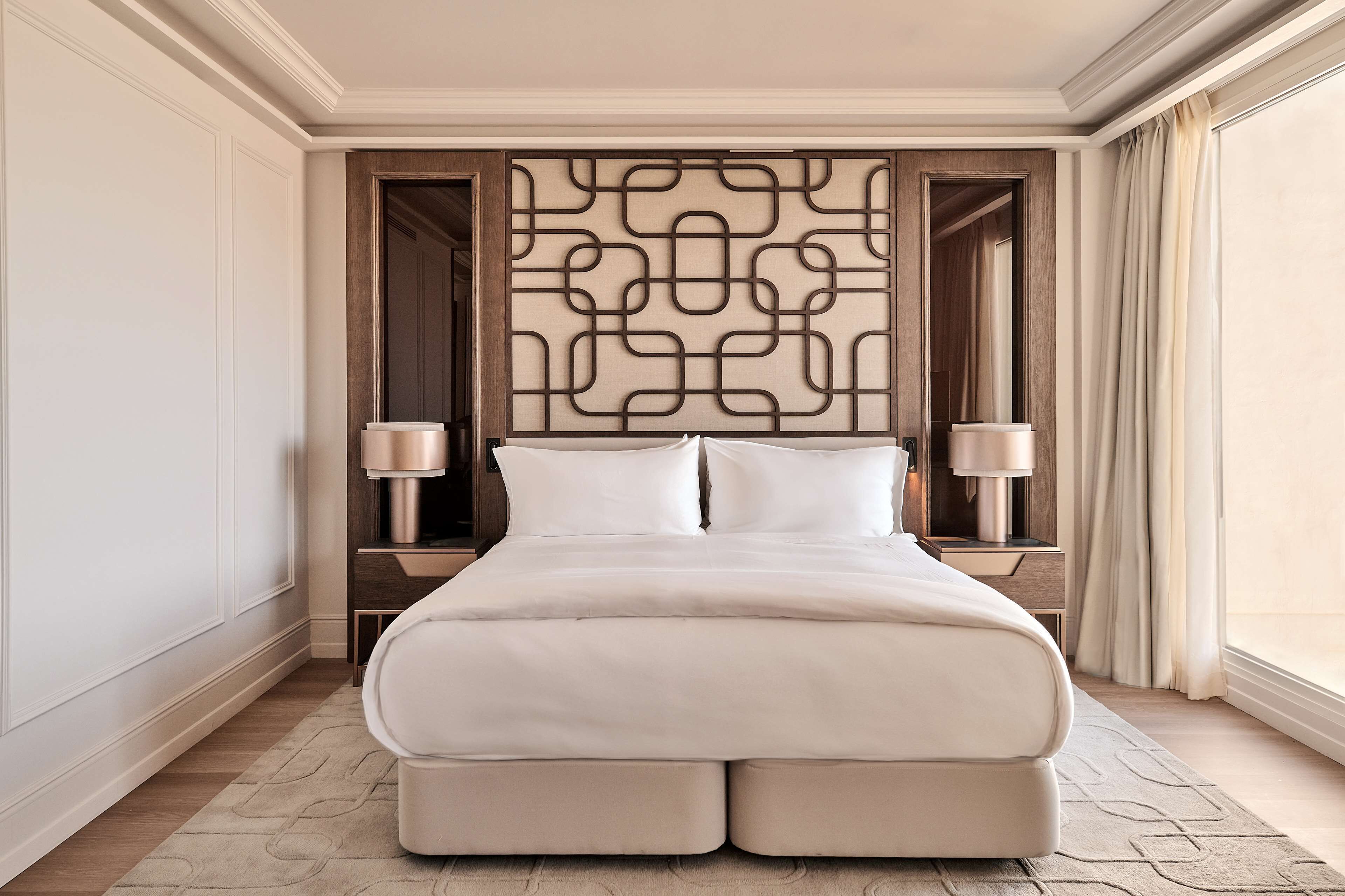 Een groot bed met twee nachtkastjes, een decoratief wandpaneel en een raam met gordijnen.