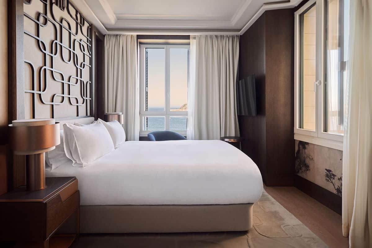 A modern hotel room with a large bed, a decorative wall panel, a television, and a view of the sea.