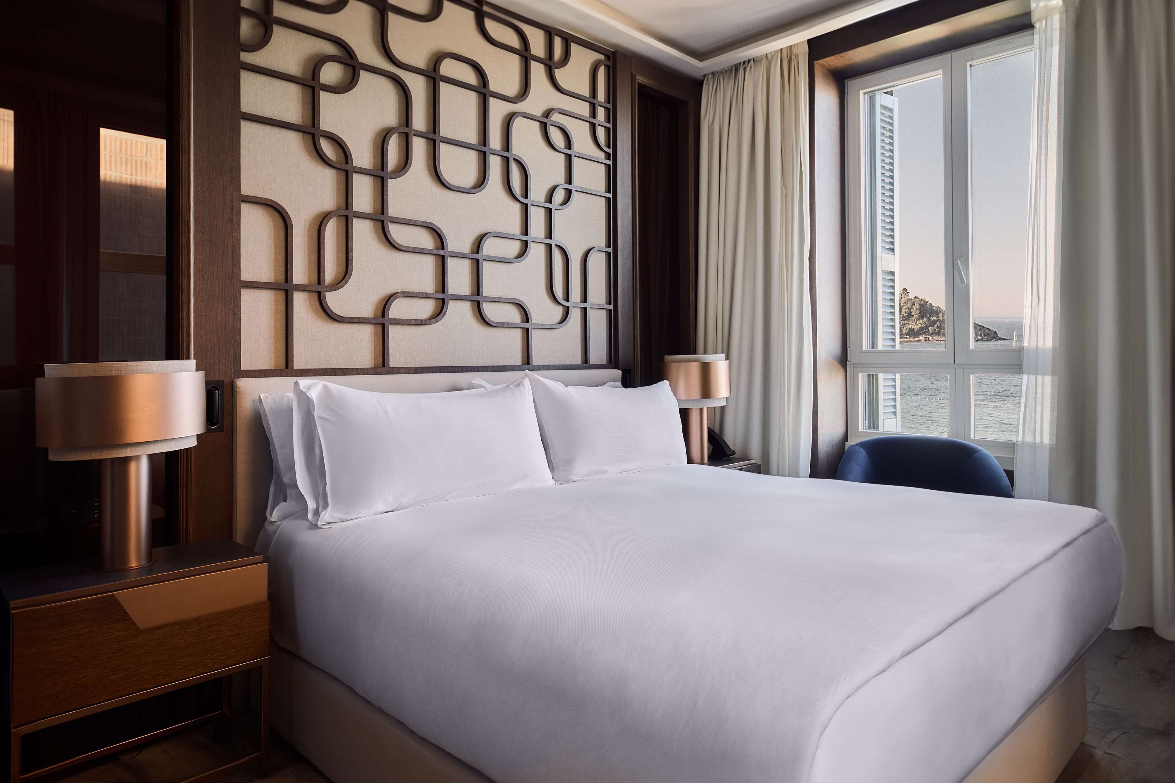 Et moderne hotelværelse med en stor seng, en dekorativ sengegavl, to sengeborde med lamper og havudsigt gennem et stort vindue.