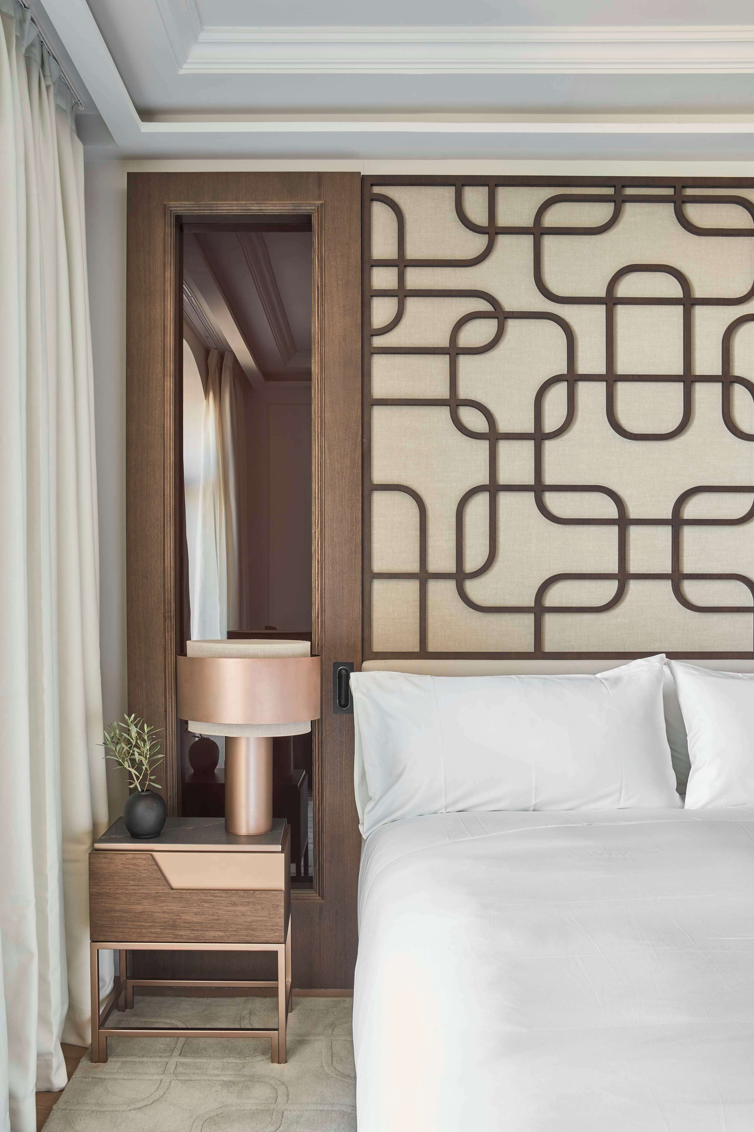 Habitación de hotel con cama, mesita de noche con lámpara, un panel decorativo en la pared y una puerta que da a otra habitación.