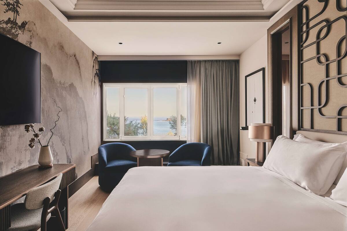 A hotel room with a large bed, two chairs, a small table, a TV, and a view of the sea through a window.