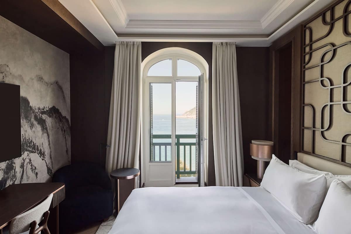 A hotel room with a large bed, a desk, a chair, and a view of the sea through an arched window.