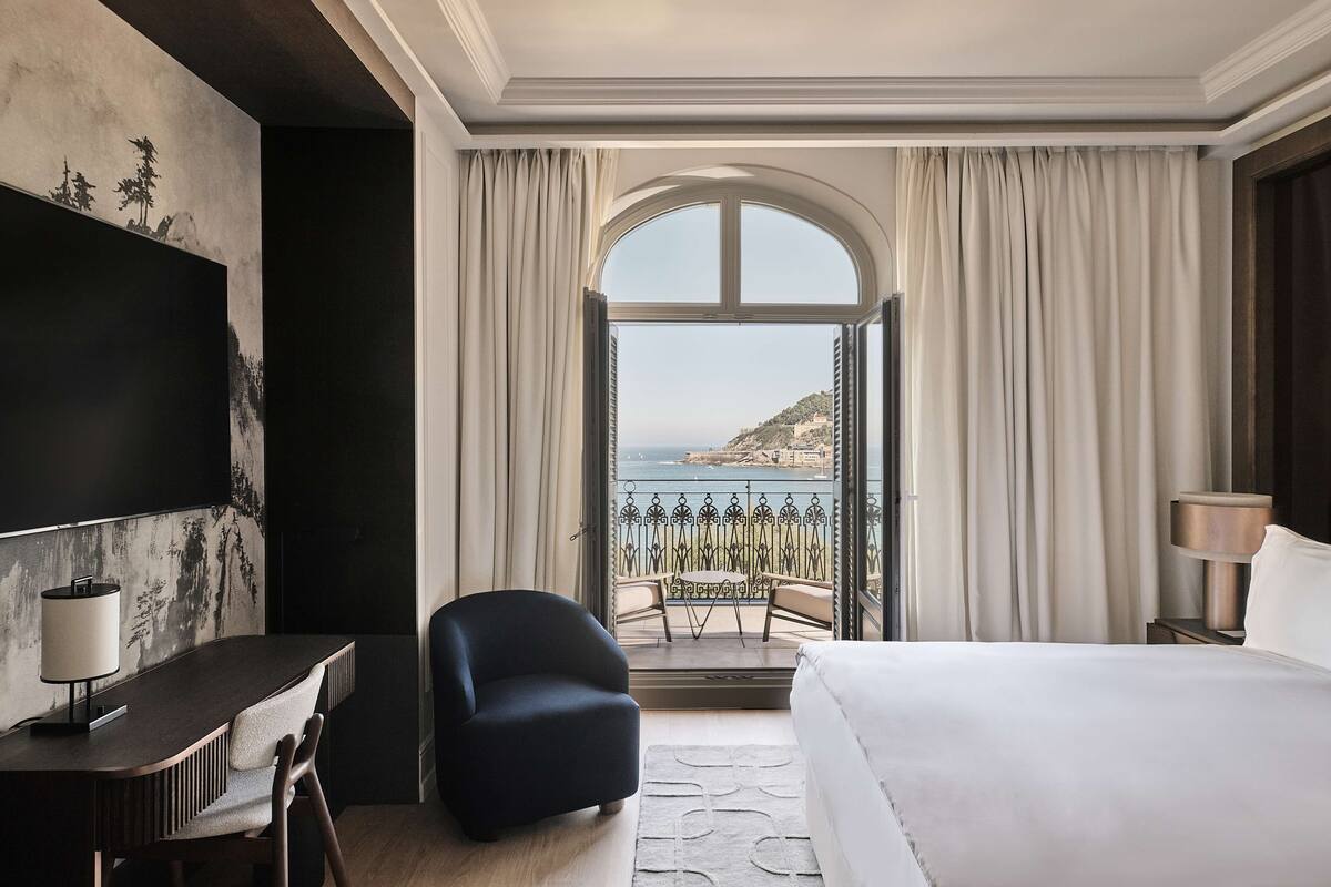 A hotel room with a bed, a desk, a chair, a TV, and a balcony with a view of the sea.