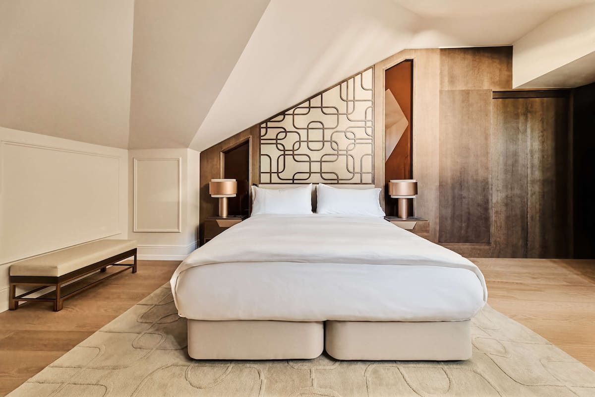 A modern bedroom with a large bed, wooden headboard, two bedside tables with lamps, a bench, and a large window.