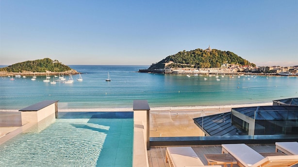 A rooftop infinity pool with clear blue water, lounge chairs, and a view of the sea with boats and a hill in the background. - Nobu Hotel San Sebastián (San Sebastián)