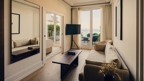 A modern living room with a sofa, a coffee table, and a TV. There is a sliding glass door leading to a balcony with a view of the sea. - Nobu Hotel San Sebastián (San Sebastián)