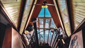 Un escalier en bois mène à une chambre située en mezzanine, d’où l’on peut admirer une maison bleue à travers une fenêtre.