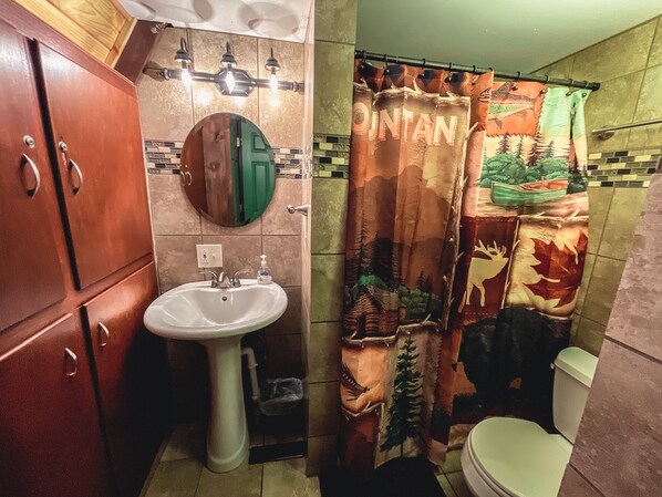 A bathroom with a wooden cabinet, a white sink, a mirror, and a shower curtain with a forest design. - Three A-frame Cabins, a provate Pond, row boats! (Rockbridge)