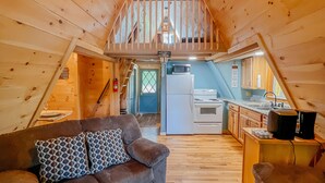 L’intérieur chaleureux d’un chalet en bois comprend une chambre à coucher située en mezzanine, une cuisine et un espace de vie avec un canapé et deux fauteuils.