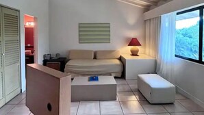 A modern hotel room with a sofa, a small table with a lamp, a window with a view of greenery, and a door leading to another room. - CACAO SUITE IN CORONADO LUXURY  CLUB & SUITES - Kitchen & golf course A (Playa Coronado)