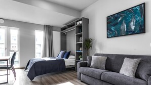 Ein modernes Schlafzimmer mit Bett, grauem Sofa, großem Fenster und an der Wand montiertem Fernseher.