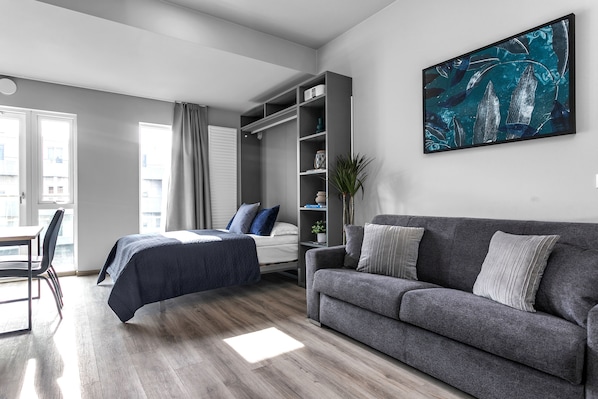 A modern bedroom with a bed, a grey sofa, a large window, and a wall-mounted TV.