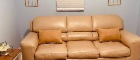 A beige leather sofa with two cushions, a wooden side table with a vase, and a patterned rug on a tiled floor.