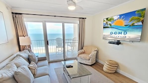 A spacious living room with a sofa, armchairs, a coffee table, and a TV displaying 'OCMD GETAWAYS'. A balcony with a view of the ocean is visible.