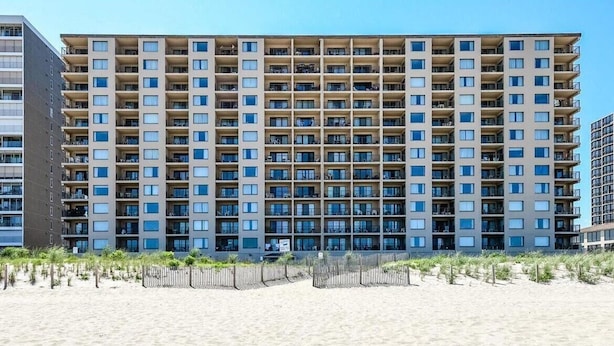 A multi-story hotel with a uniform facade, balconies, and a sandy beach in the foreground.