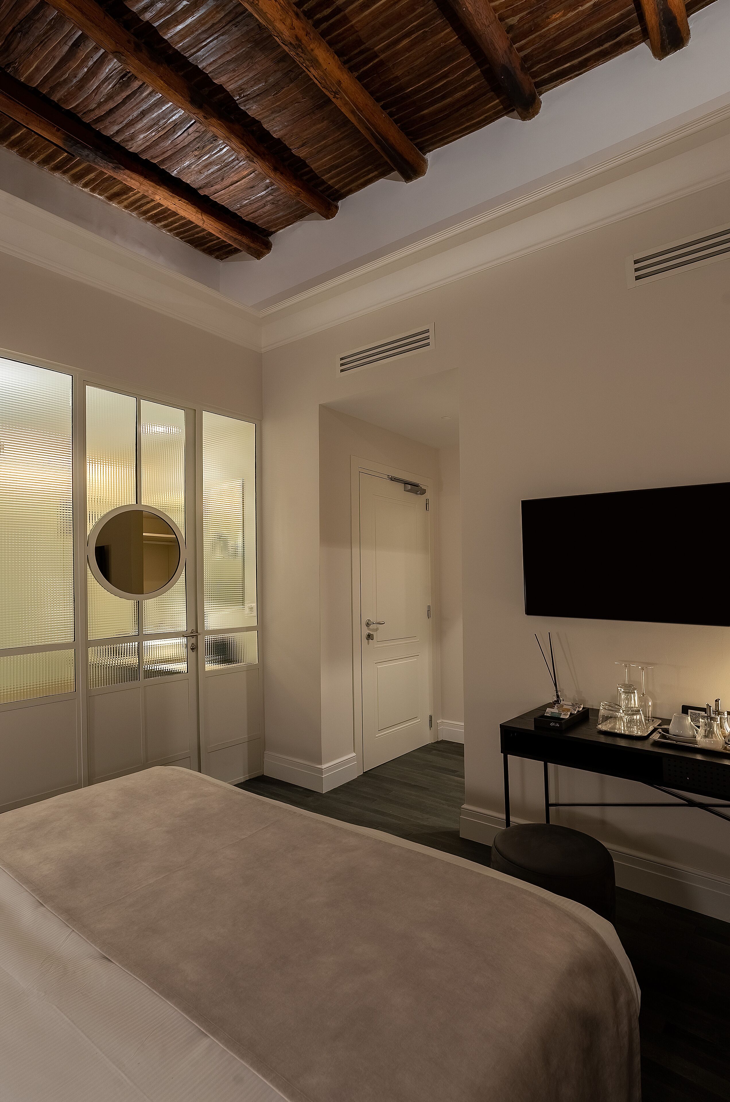 A hotel room with a large bed, a desk with a flat-screen TV, a round mirror, and a wardrobe with frosted glass doors.