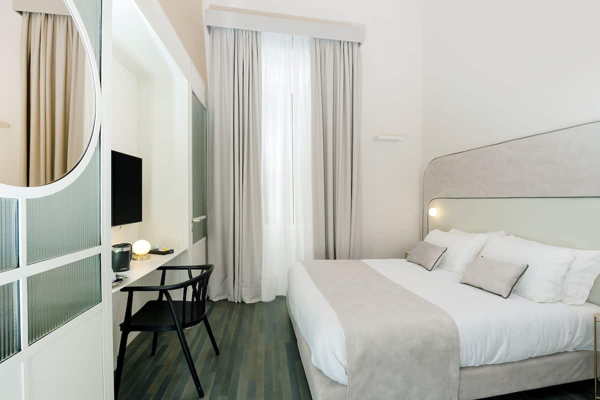 A hotel room with a bed, a desk with a chair, a mirror, and a television.
