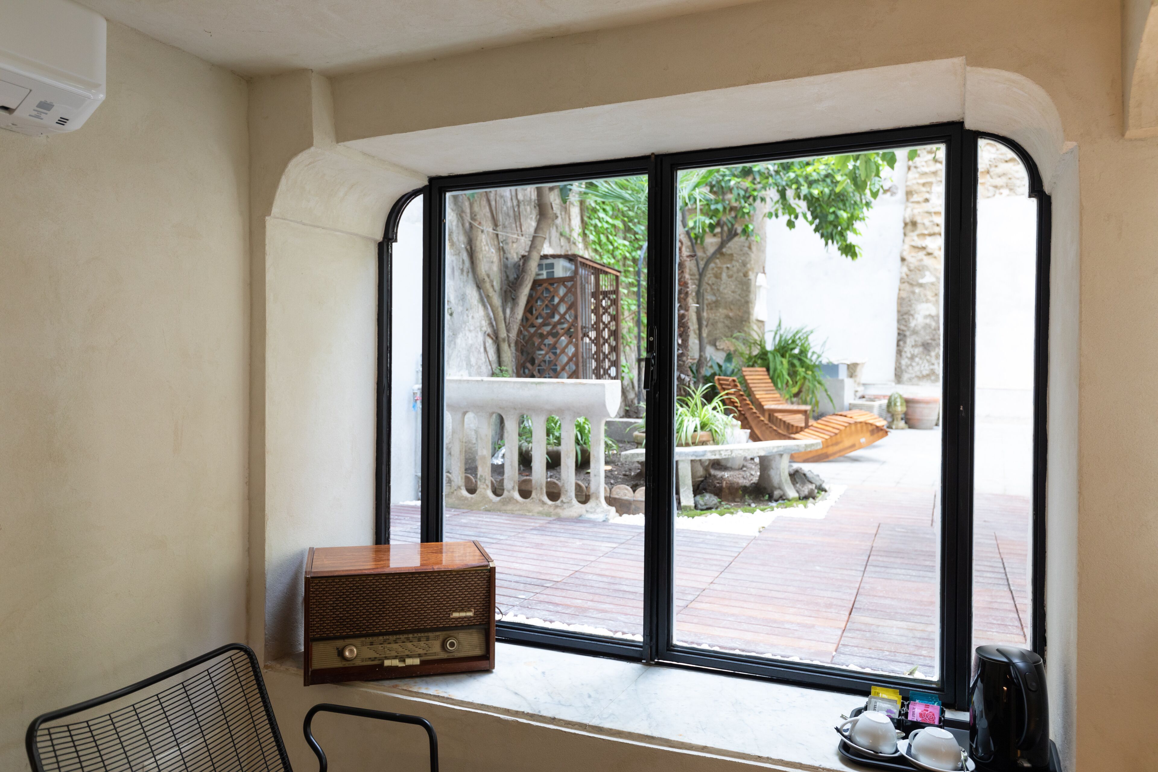Una stanza con una grande finestra che si affaccia su un patio con ringhiera bianca, una panca di legno e piante in vaso. Una radio vintage è posata sul davanzale.