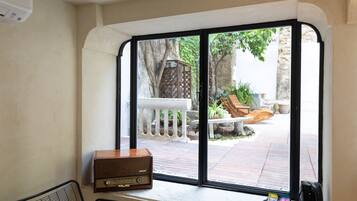 Una stanza con una grande finestra che si affaccia su un patio con ringhiera bianca, una panca di legno e piante in vaso. Una radio vintage è posata sul davanzale.