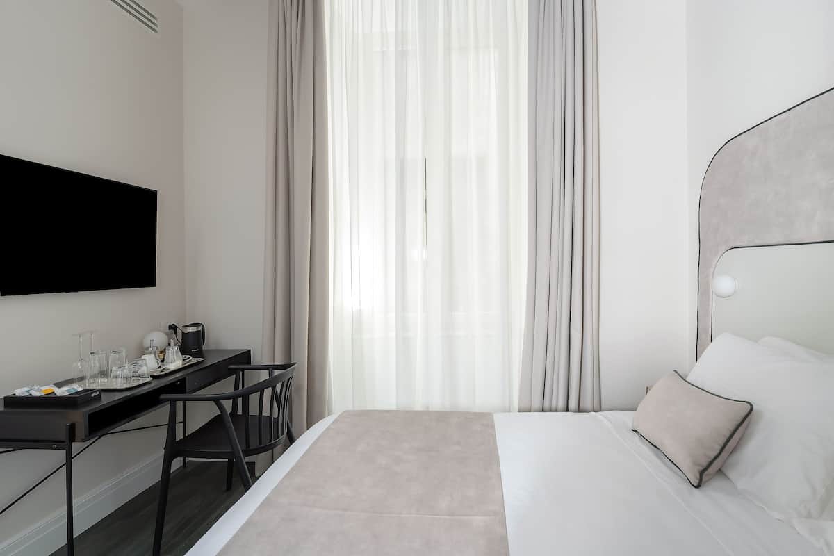 A modern hotel room with a bed, a desk with a chair, a flat-screen TV, and a large window with sheer curtains.