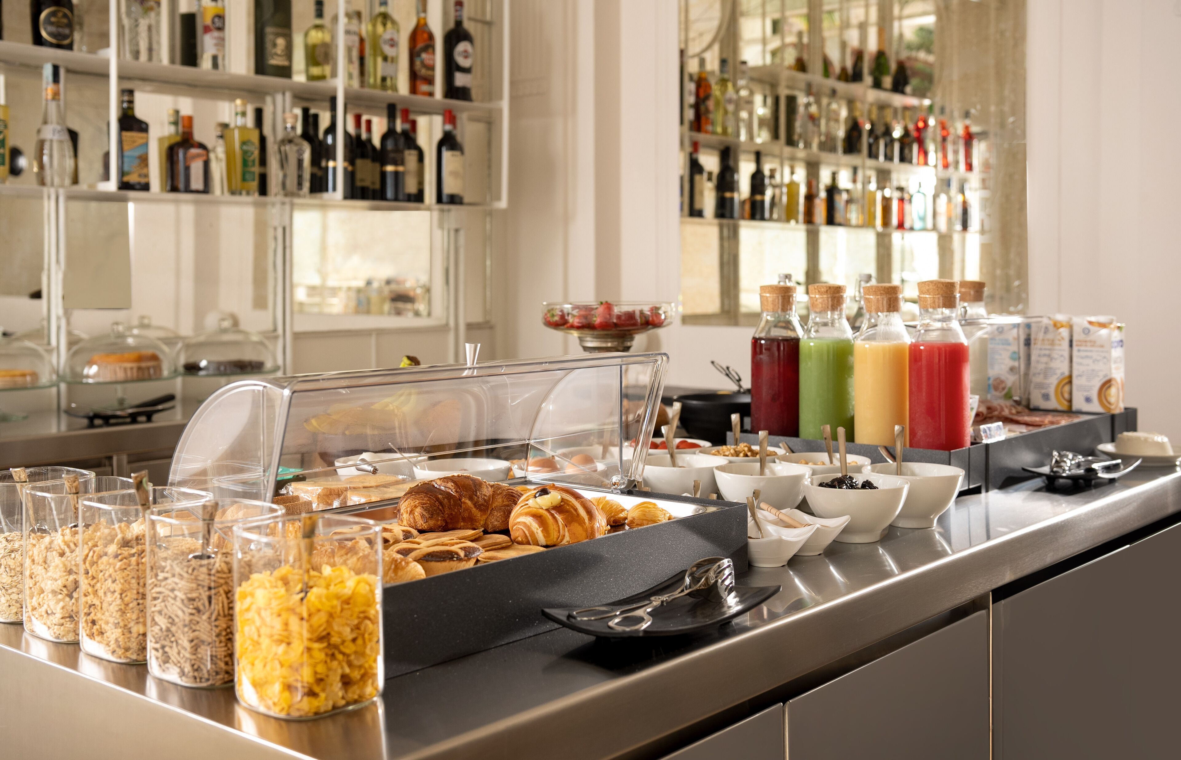 Un bufé de desayuno con diversos alimentos y bebidas, incluyendo botes de cereales, croissants y bebidas de colores.