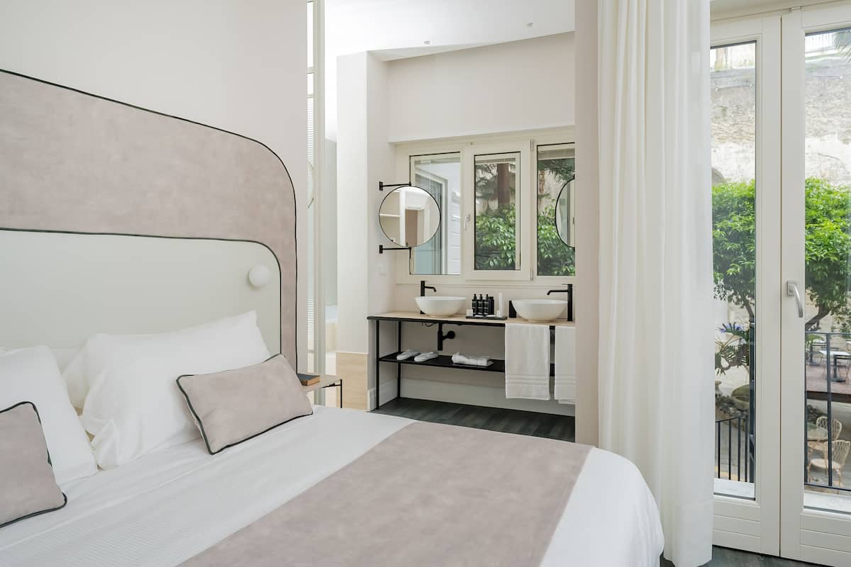 A bedroom with a bed, a bathroom with a sink and mirror, and a view of greenery outside.