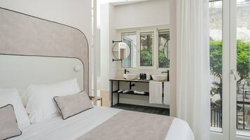Ein Schlafzimmer mit Bett, ein Badezimmer mit Waschbecken und Spiegel sowie Blick auf Grünflächen.