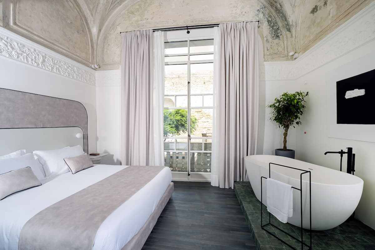 A modern bedroom with a large bed, a freestanding bathtub, and a balcony with curtains.