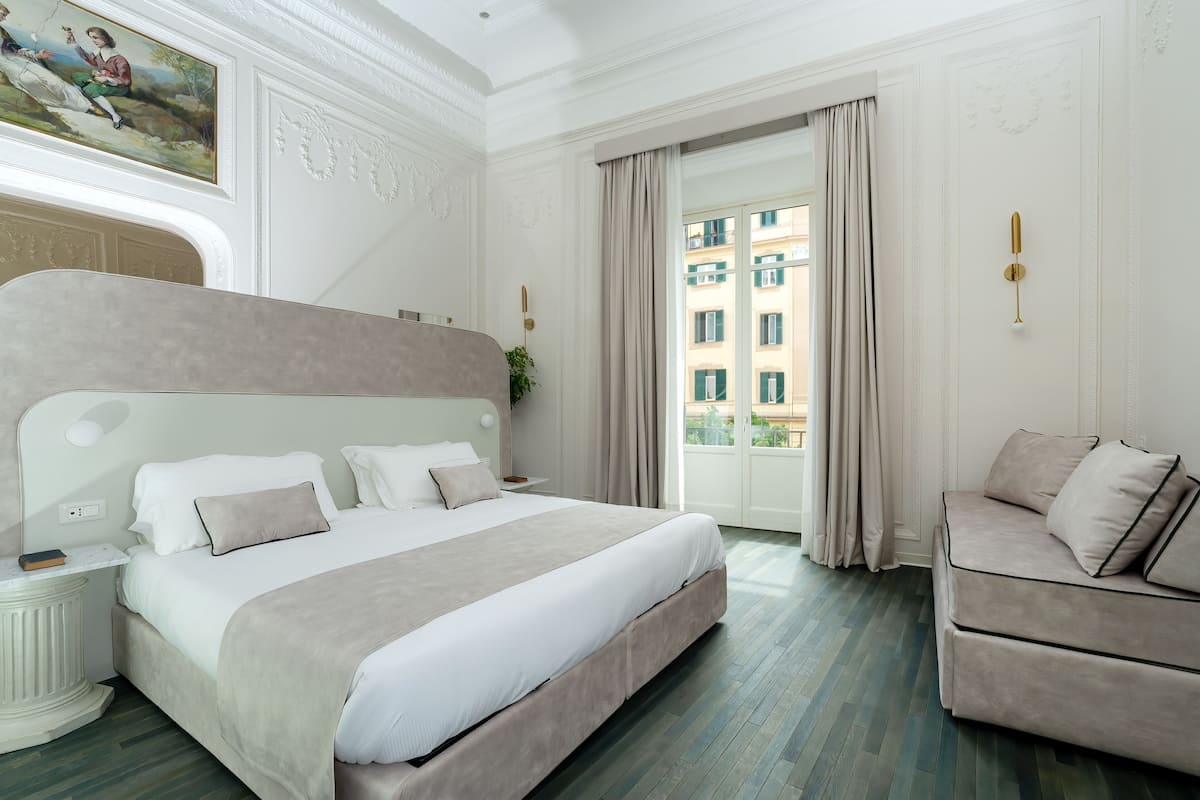 A hotel room with a large bed, a sofa, a painting on the wall, and a view of buildings through the window.